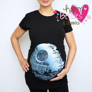Pode incluir: Uma camiseta de maternidade preta com um gráfico azul e branco da Estrela da Morte. O texto "happy Bellies & babies" está impresso em rosa, marrom e verde no canto superior direito da camiseta.