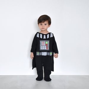 May include: A young child dressed in a black Darth Vader costume with a cape. The costume features a printed image of Darth Vader's chest panel and belt.