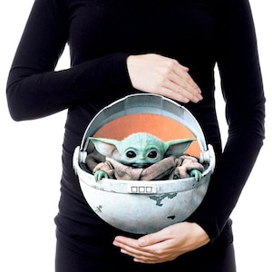 May include: A black maternity top featuring a graphic of a green alien character in a floating pod. The alien is wearing a brown robe and has large ears.