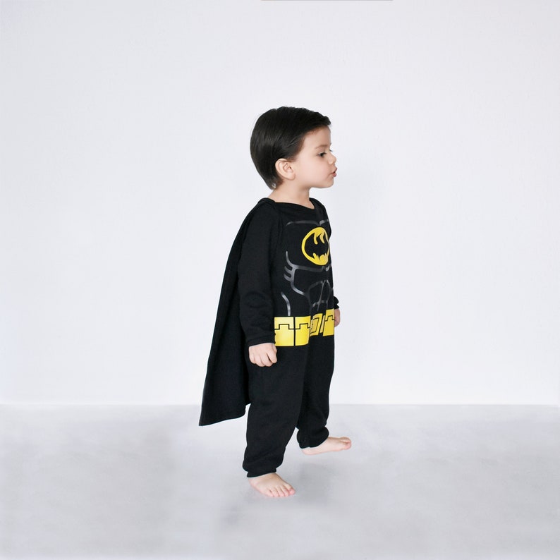 May include: A child wearing a black Batman costume with a yellow belt and cape. The costume features a printed image of Batman's chest and a bat symbol.