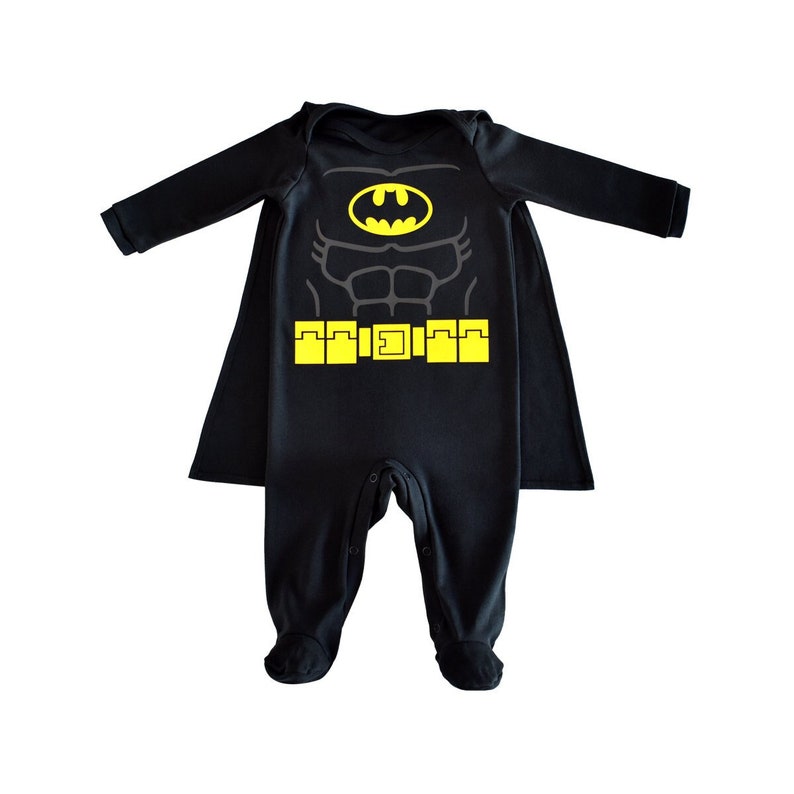 May include: Black baby onesie with a yellow Batman logo and a matching cape. The onesie features a printed image of Batman's torso with a yellow belt.