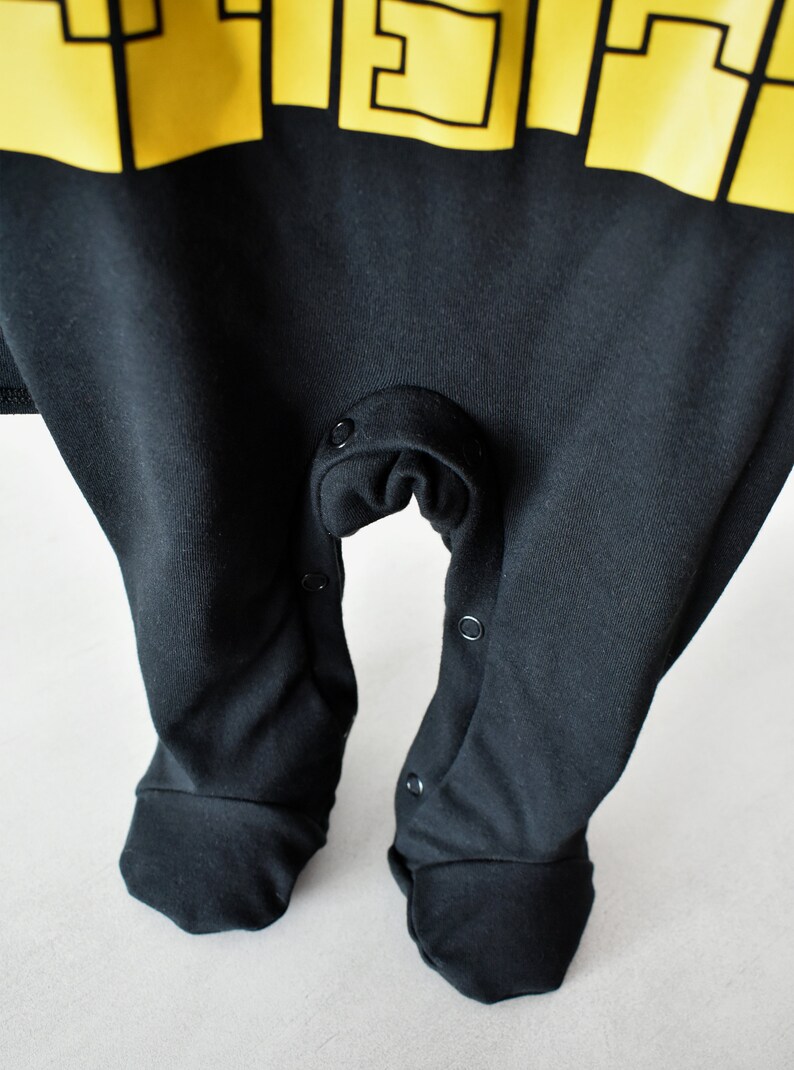 May include: Black baby onesie with yellow lettering that says "F.I.E.S.T.A." The onesie has snaps at the crotch and on the legs.
