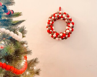 以下が含まれることがあります： 白い壁に赤い、白い、そして灰色のフェルトボールのクリスマスリースが掛かっています。リースは、円形に縫い合わされたフェルトボールで作られています。