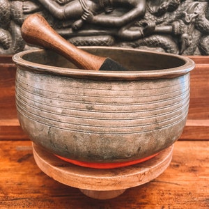 Puede incluir: Un cuenco de canto plateado con un mazo de madera sobre un soporte de madera con un cojín rojo. El cuenco tiene una superficie texturizada y está decorado con líneas horizontales.