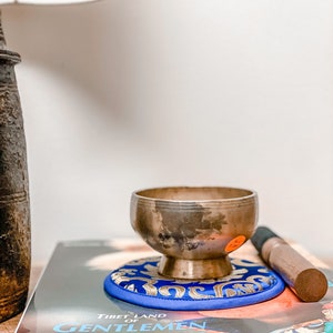 Pode incluir: Uma tigela de canto de bronze repousa sobre uma almofada azul e dourada em cima de um livro intitulado "Tibet Land of Gentlemen Brigands". Um malho de madeira está ao lado da tigela. Uma lâmpada ilumina a cena.