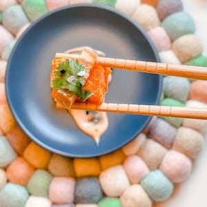 May include: A close-up of a salmon and avocado poke bowl with a creamy sauce, garnished with cilantro and sesame seeds. The bowl is on a colorful handmade trivet.