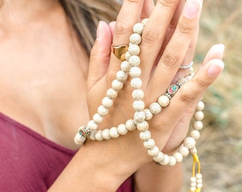 May include: A person is holding a white mala bead necklace with a silver charm. The beads are small and round, and the charm is a small, ornate design. The person's hands are clasped together, and the necklace is draped over their fingers.