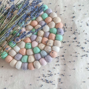 May include: A round, multi-colored felt coaster with a lavender sprig on top. The coaster is made of individual felt balls in shades of pink, green, beige, and purple.