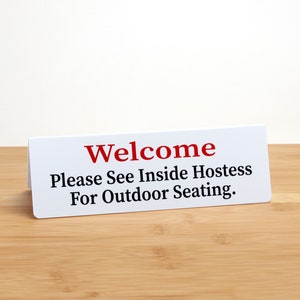 May include: White sign with red text that says "Welcome" and "Please See Inside Hostess For Outdoor Seating."