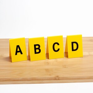 May include: Four yellow rectangular markers with black capital letters A, B, C, and D printed on them. The markers are standing upright on a light brown wooden surface, against a white background.