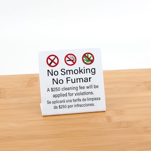 May include: A white sign with red no-smoking symbols and the text "No Smoking No Fumar." The sign also states that a $250 cleaning fee will be applied for violations. The sign is on a wooden surface.