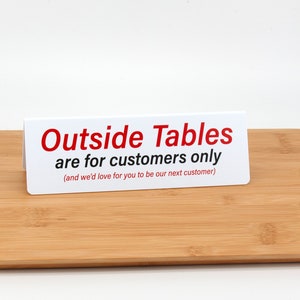 May include: White sign with red text that reads "Outside Tables are for customers only (and we'd love for you to be our next customer)"