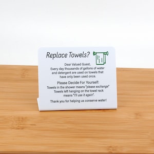 May include: White sign with green text that reads "Replace Towels?" and includes a small illustration of a towel hanging on a rack. The sign explains that towels in the shower mean "please exchange" and towels left hanging on the rack mean "I'll use it again".