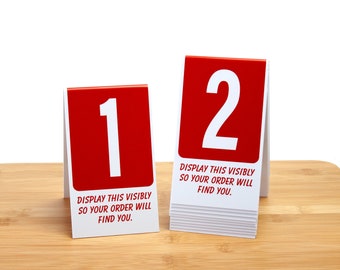May include: Two red table number signs with white numbers. The sign on the left is number 1 and the sign on the right is number 2. Both signs have the text "DISPLAY THIS VISIBLY SO YOUR ORDER WILL FIND YOU."