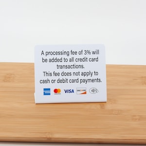 May include: A white sign with text about a 3% processing fee for credit card transactions. The sign includes logos for American Express, Mastercard, Visa, Discover, and a contactless payment symbol. The sign is on a light brown wooden surface.