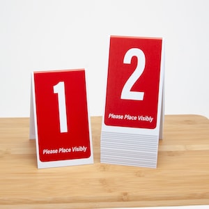 May include: Two red table number signs with white numbers and the text "Please Place Visibly". The number signs are on a wooden surface. The number signs are 1 and 2.