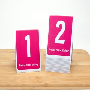 May include: Two pink table number signs with white numbers and the text "Please Place Visibly". The number one sign is on the left and the number two sign is on the right.
