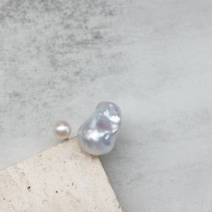 May include: Two pearls, one small and round, the other large and baroque, rest on a light-colored surface against a gray background. The baroque pearl has an irregular shape and a lustrous, iridescent surface.