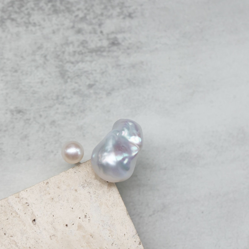 May include: Two pearls, one small and round, the other large and baroque, rest on a light-colored surface against a gray background. The baroque pearl has an irregular shape and a lustrous, iridescent surface.