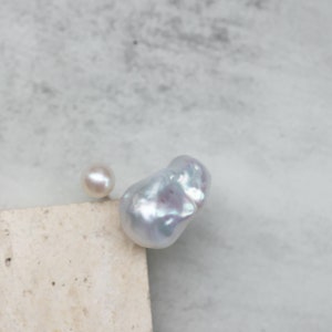 May include: Two pearls, one round and one baroque, rest on a neutral-toned surface against a soft, blurred background. The baroque pearl has an irregular shape and a lustrous, iridescent surface. The round pearl is smaller and has a smooth, creamy appearance.