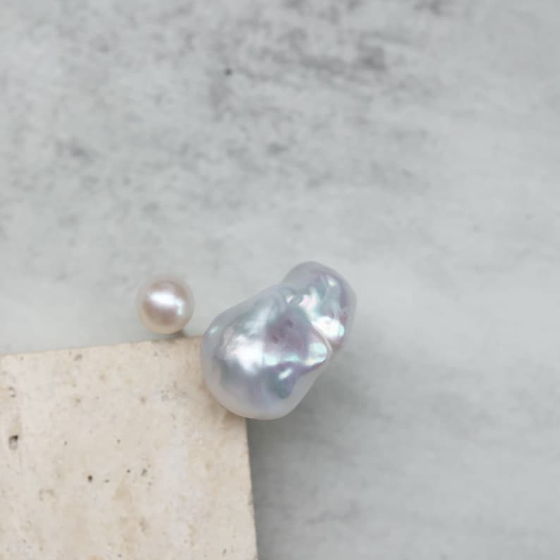 May include: Two pearls, one round and one baroque, rest on a neutral-toned surface against a soft, blurred background. The baroque pearl has an irregular shape and a lustrous, iridescent surface. The round pearl is smaller and has a smooth, creamy appearance.