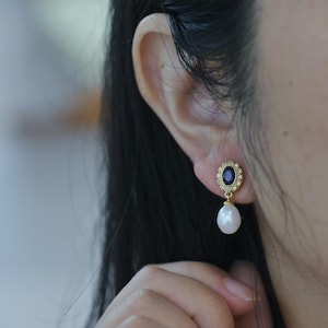 Peut inclure: Boucles d'oreilles en or avec une pierre ovale bleue entourée de petites pierres claires et une perle blanche pendante.