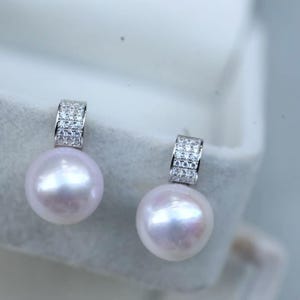 Op de afbeelding: Een paar elegante oorbellen met grote, glanzende, lichtroze parels. Elke oorbel heeft een zilverkleurige, rechthoekige bovenkant bezet met kleine, sprankelende, heldere steentjes. De oorbellen zijn ontworpen om te bungelen.
