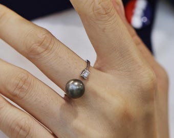 Könnte beinhalten: Ein silberner Ring mit einer dunkelgrauen Perle und einem kleinen quadratischen Diamanten. Der Ring befindet sich an einem Finger und hebt den Kontrast zwischen dem Metall und den Edelsteinen hervor. Modernes und elegantes Design.