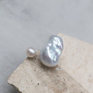 May include: Two lustrous pearls rest on a textured, light-colored surface. One is a small, round pearl, while the other is a large, irregularly shaped baroque pearl with iridescent highlights, reflecting a spectrum of colors.