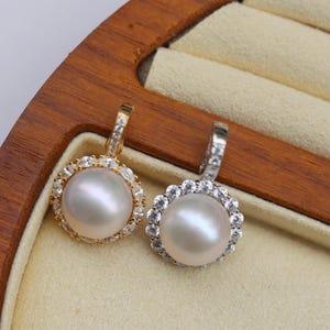 Könnte beinhalten: Zwei Perlenanhänger mit einem Heiligenschein aus kleinen, klaren Steinen. Der Anhänger links hat eine goldene Fassung, der Anhänger rechts eine silberne Fassung.