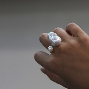 May include: A unique ring featuring two lustrous, irregularly shaped white pearls. One large pearl sits atop the finger, while a smaller pearl is positioned below. The ring's design highlights the natural beauty of the pearls, creating a statement piece.