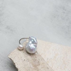 May include: A silver ring featuring two pearls. One is a small, round pearl, and the other is a large, irregularly shaped baroque pearl. The ring is displayed on a light-colored, textured surface. Jewelry.