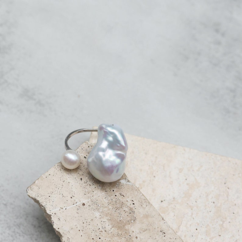May include: A silver ring featuring two pearls. One is a small, round pearl, and the other is a large, irregularly shaped baroque pearl. The ring is displayed on a light-colored, textured surface. Jewelry.