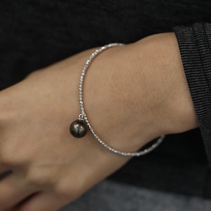 Puede incluir: Un brazalete plateado con un solo dije de perla negra. El brazalete está hecho de pequeñas cuentas redondas.