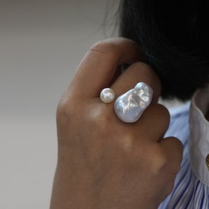 May include: A unique ring featuring two pearls. One is a small, round pearl, and the other is a large, irregularly shaped baroque pearl. The pearls are white and have a lustrous sheen. The ring is a statement piece of jewelry.