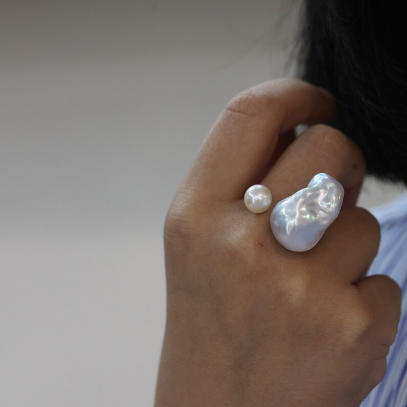 May include: A unique ring featuring two lustrous pearls. One is a small, round pearl, and the other is a large, irregularly shaped baroque pearl. The pearls have a creamy white color and a shimmering surface. The ring is a statement piece of jewelry.