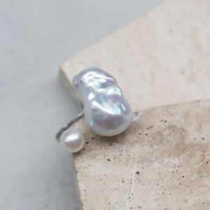 May include: A silver ring featuring a large, irregular-shaped white pearl and a smaller, round pearl. The pearls have a lustrous, iridescent sheen. The ring is set against a neutral, textured background.