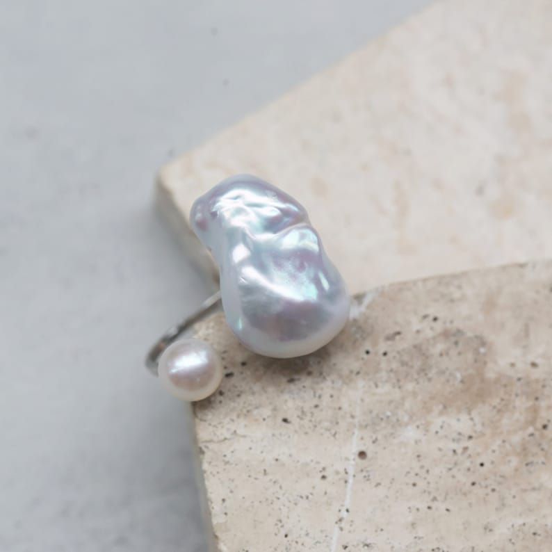 May include: A silver ring featuring a large, irregular-shaped white pearl and a smaller, round pearl. The pearls have a lustrous, iridescent sheen. The ring is set against a neutral, textured background.