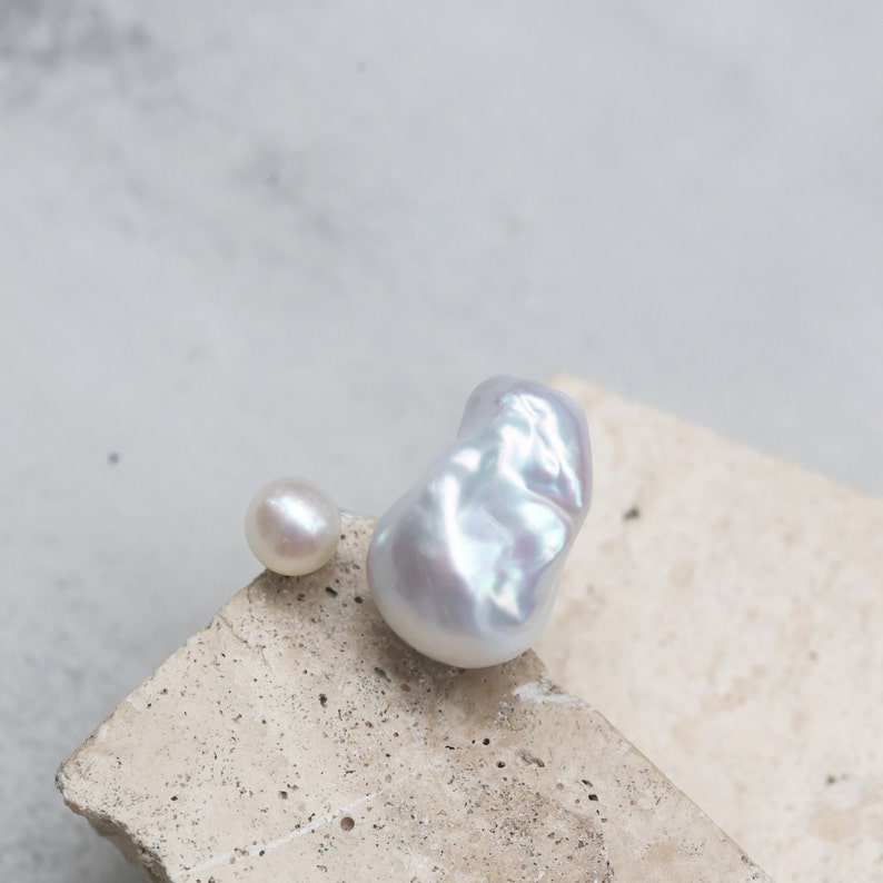 May include: Two pearls, one small and round, the other large and baroque, rest on a textured, light-colored surface. The larger pearl has a lustrous, iridescent sheen, reflecting shades of white and pale blue. The smaller pearl is a soft, creamy white.
