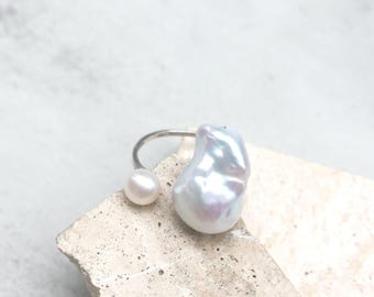 May include: A silver-toned open ring featuring two pearls of different sizes. One end of the band holds a small, round, white pearl, while the other end features a large, irregular, baroque-style pearl with a lustrous, iridescent white finish.