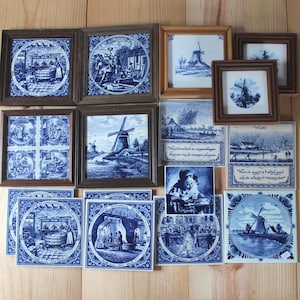 Op de afbeelding: Een verzameling vierkante keramische tegels met blauw-witte illustraties van Nederlandse landschappen, windmolens en historische scènes. Sommige tegels zijn ingelijst in hout, andere zijn niet ingelijst. Verschillende tegels bevatten Nederlandse teksten.