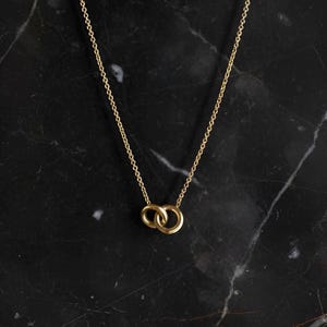 May include: A gold-toned necklace featuring a delicate chain and a pendant composed of two small, interlocking circular rings. The metal has a smooth, polished finish, and the design is simple, consisting of the chain and the linked rings.
