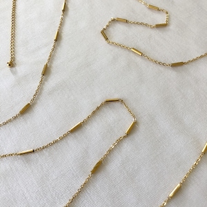 Könnte beinhalten: Eine goldene Kettenkette mit einer zarten Kette und kleinen, rechteckigen goldenen Perlen. Die Kette ist verstellbar und kann in verschiedenen Längen getragen werden.