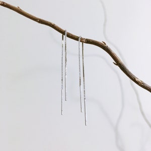 May include: Silver threader earrings suspended from a brown branch against a white backdrop. The earrings feature a fine chain and a small, elongated bar. The design is minimalist and elegant, suitable for everyday wear.