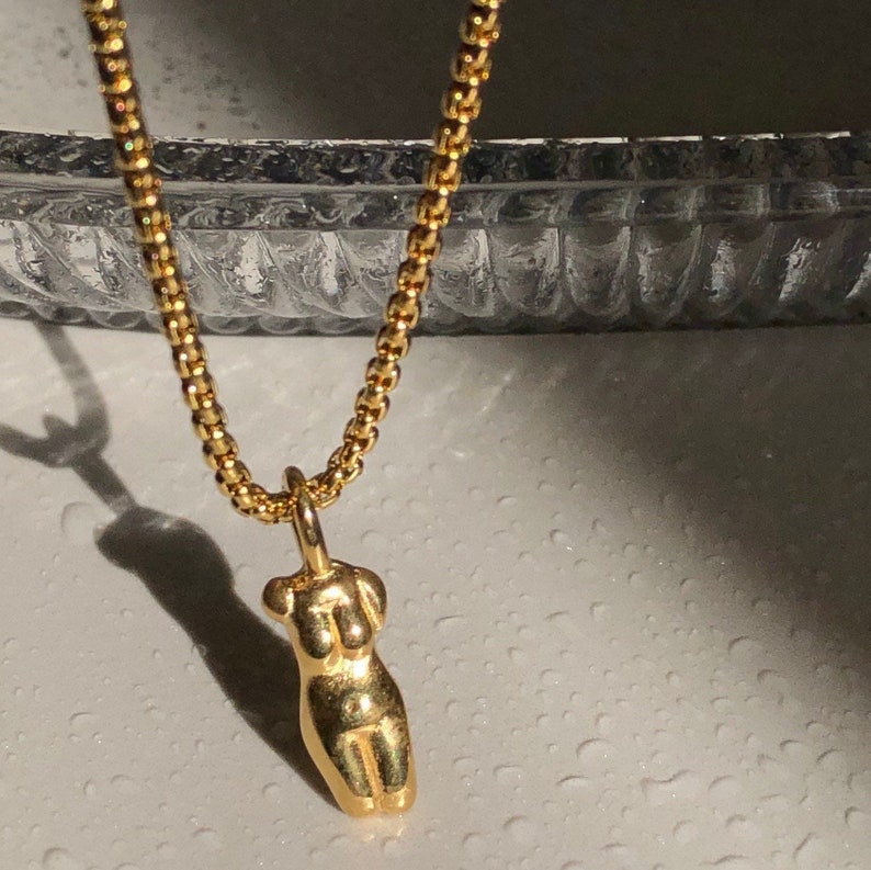 May include: A gold-toned necklace featuring a box chain and a pendant shaped like a stylized human torso. The pendant has a smooth, reflective surface and is suspended from the center of the chain, which has a repeating square link pattern.