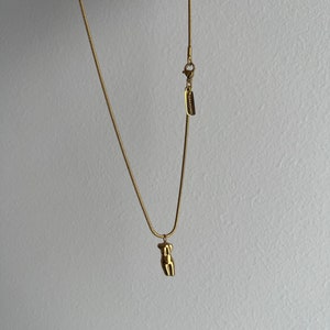 May include: A gold-toned snake chain necklace featuring a small, polished pendant shaped like a human torso. The necklace includes a lobster claw clasp and a rectangular metal tag engraved with the text SURADISIRES near the closure.