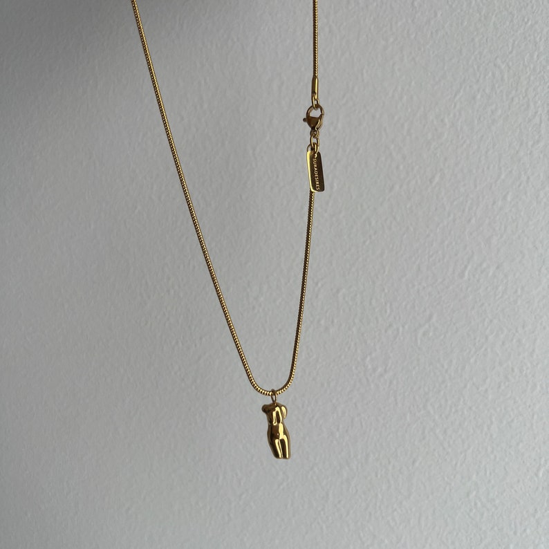 May include: A gold-toned snake chain necklace featuring a small, polished pendant shaped like a human torso. The necklace includes a lobster claw clasp and a rectangular metal tag engraved with the text SURADISIRES near the closure.