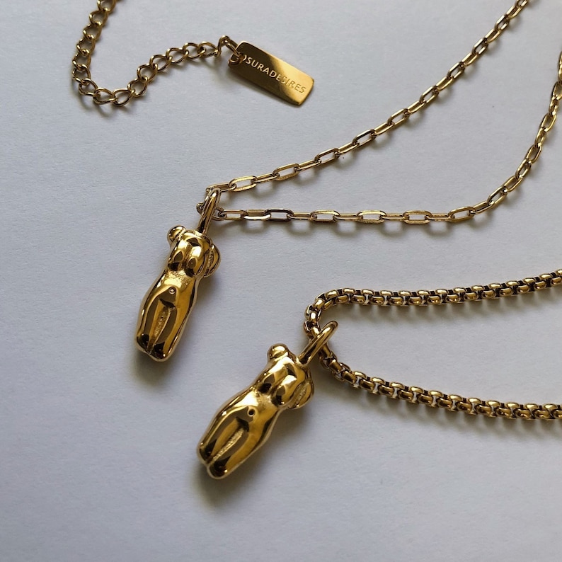 May include: Two gold-toned necklaces featuring polished pendants shaped like human torsos. Each pendant hangs from a metallic chain. One chain is a flat link style, the other is a rounded box chain. A small rectangular tag is engraved with SURADESIRES.