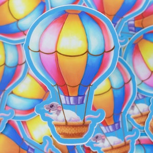 May include: A colorful hot air balloon with a white rat riding in the basket. The balloon is pink, yellow, blue, and orange. The rat is wearing a purple hat.