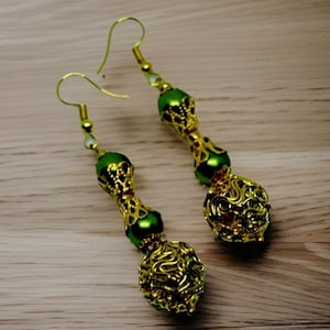 Puede incluir: Un par de pendientes colgantes dorados con detalles verdes. Los pendientes presentan un diseño de filigrana dorada y cuentas verdes. Los pendientes tienen un cierre de gancho y se exhiben sobre una superficie de madera marrón claro.